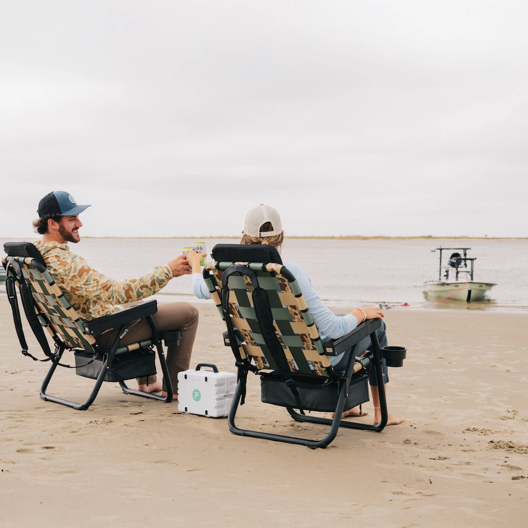 Two friends relaxing on the beach in PARKIT × Free Fly Eclipse Recliners in Vintage Camo, with a Turtlebox speaker playing between them and a skiff anchored in the background. These premium folding outdoor chairs feature a built-in cooler, backpack straps, and recline function perfect for beach days, boat trips, or backyard hangs for dads, moms, and outdoor enthusiasts alike.