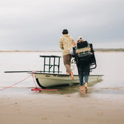 Two people walking barefoot toward a skiff, one carrying the PARKIT × Free Fly Eclipse Recliner in Vintage Camo using its backpack carry straps. The folding outdoor chair features an integrated cooler and rugged aluminum frame the ultimate portable beach, boat, and camping chair for couples, adventurers, and weekend explorers.