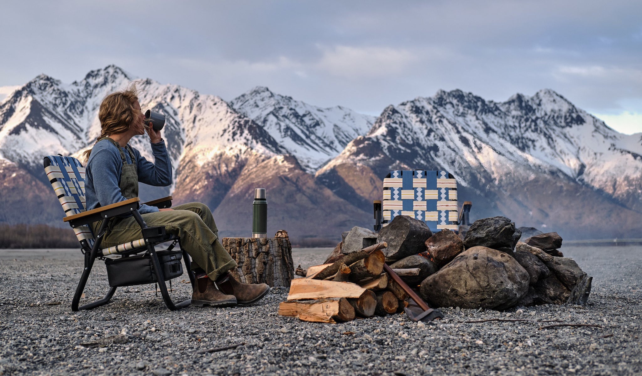 Women Sitting in PARKIT voyager outdoor chair in Alaska with a beautiful mountain beackground and the makings of a campfire in the foreground. She is drinking from a canteen while sitting in the chair.