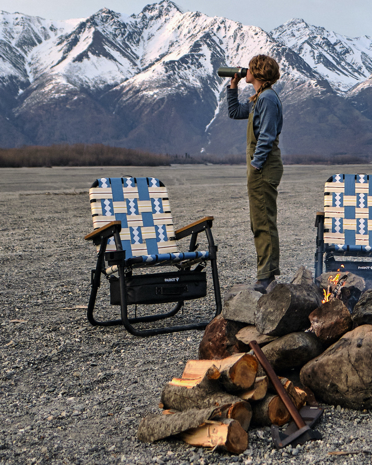 Woman standing infrom of campfire set up with her Voyager outdoor chairs in Alaska with a beatiful mountain range in the background.