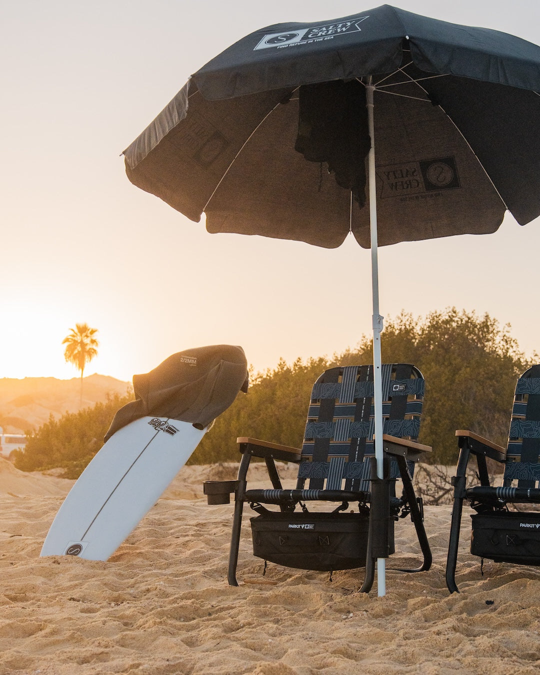 PARKIT and Salty Crew Collaboration Voyager beach boat and camping Chair with boat safe rubber feet, cooler, fishing rod accessory, cupholder, and overlander carry straps sitting on the beach on a perfect summer sunset after a day of surfing, fishing and chilling at the beach with an umbrella covering the two chairs with a palm tree in the background.