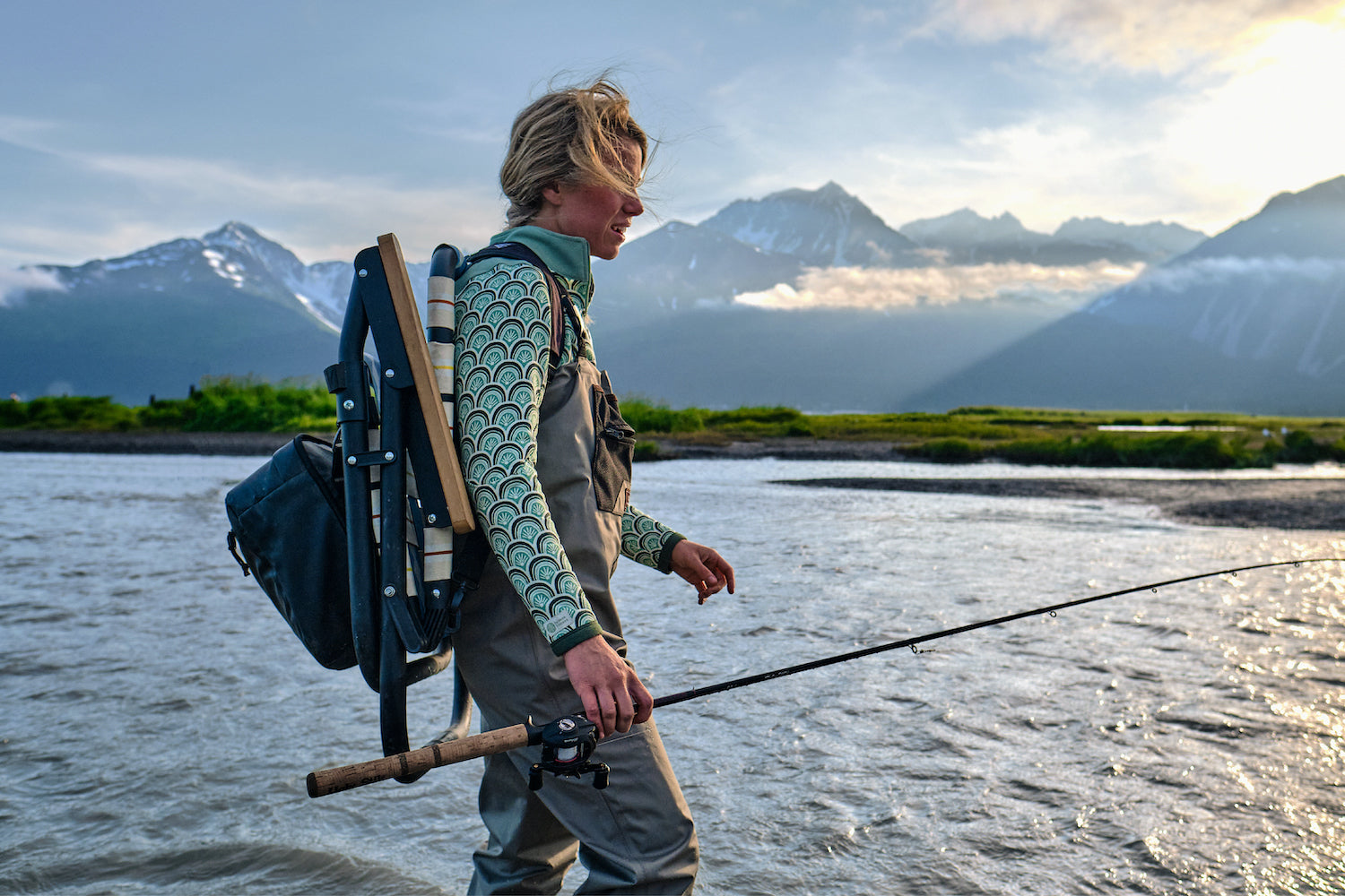 A woman walking through the river in Alaska wearing the Voyager Outdoor Chair as a Backpack while she works the river with her fly fishing rod.
