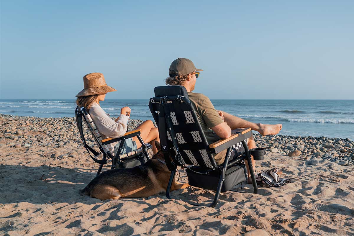 Foldable reclining lawn chair set up on the beach during sunset with guy, girl, and dog looking out to the ocean