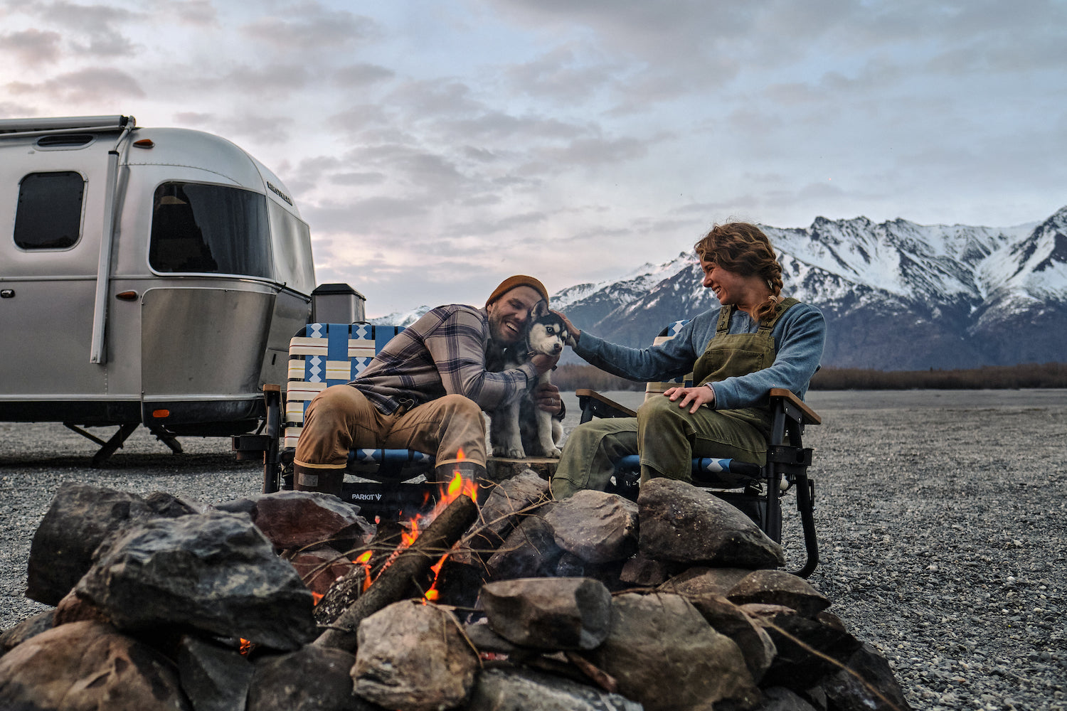 A husband and wife with sitting around the campfire with their dog while sitting in the Voyager outdoor chairs from PARKIT with their AirStream and Alaska Mountains in the background.