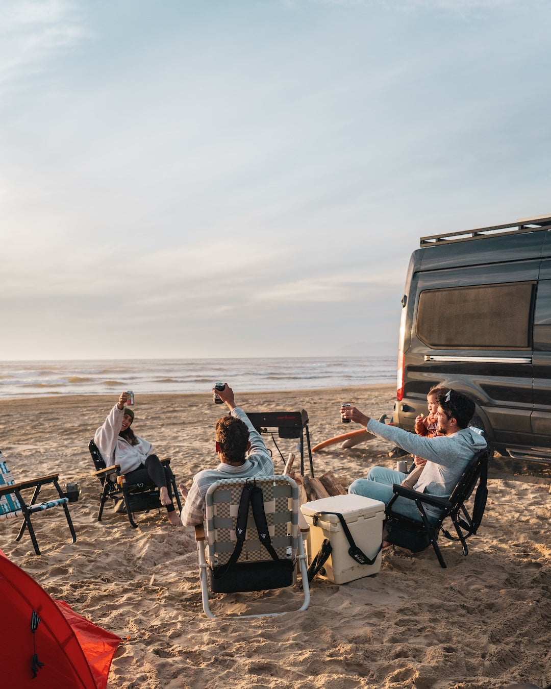 Friends sitting in PARKIT Voyager beach chairs at their camping site in Pismo Beach where their camper van is parked on the sand and the sun sets with crashing waves in the background.