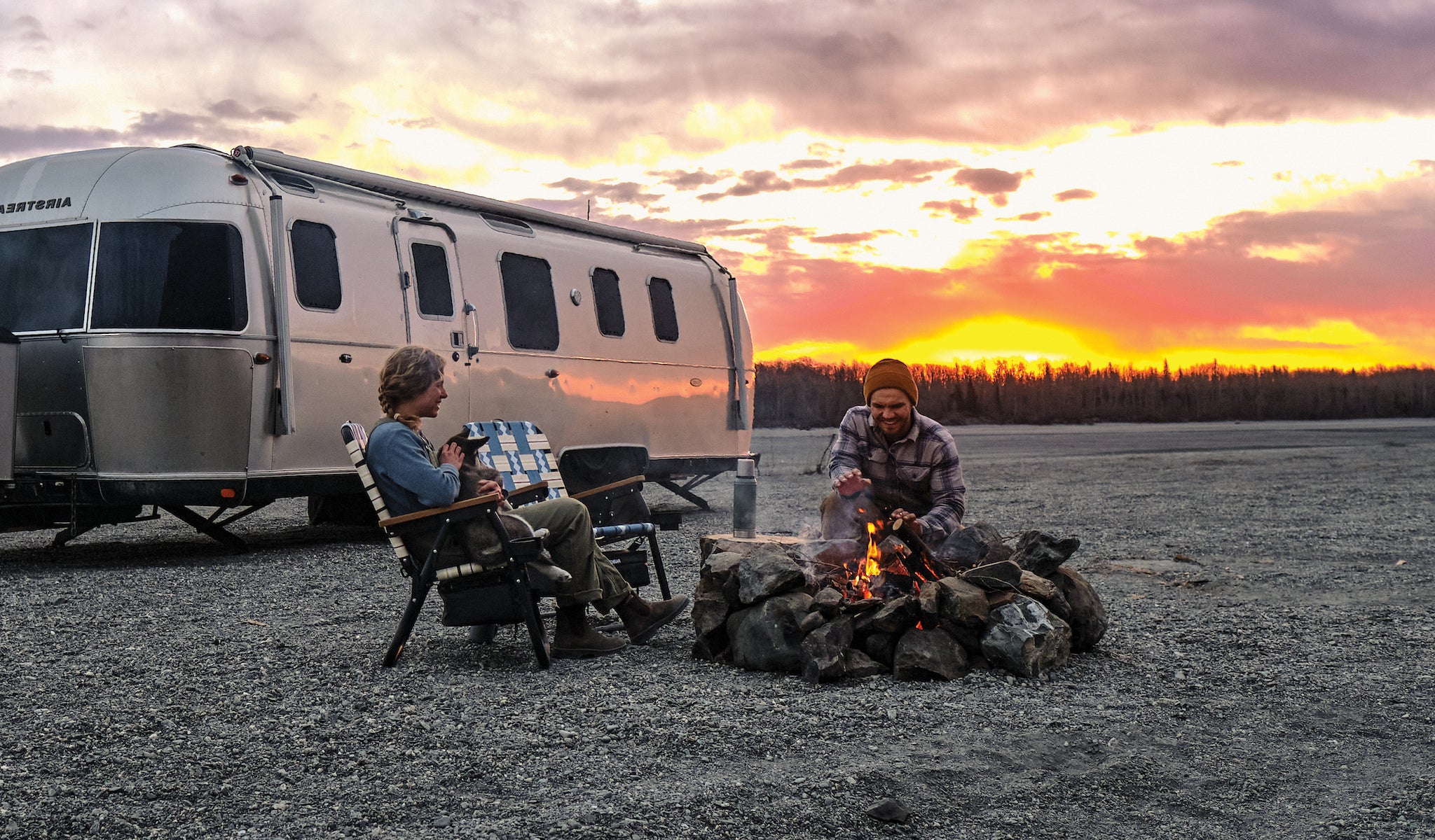 A couple sitting around the fire in their parkit voyager chairs while the sun sets and their airstream camper is in the background.