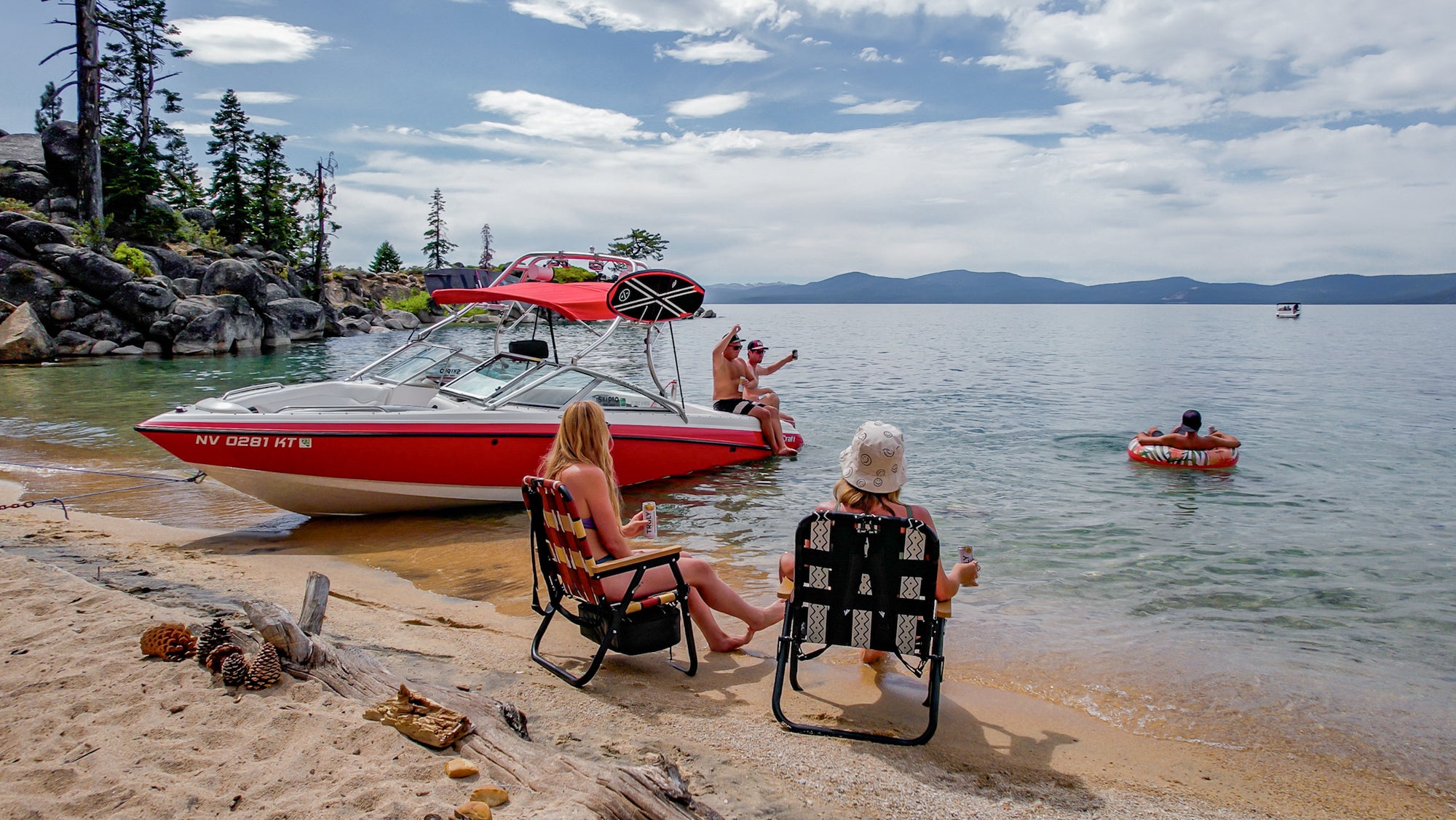 a group of people at lake Tahoe with their boat pulled up on the shore, people in the water and two people in beach chairs