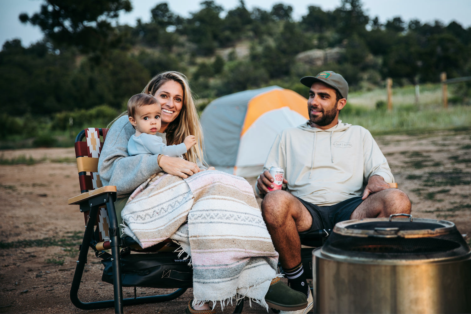 A Husbad and wife sitting around the fire at their campsite with their Baby Boy while they enjoy their Voyager outdoor chairs by PARKIT