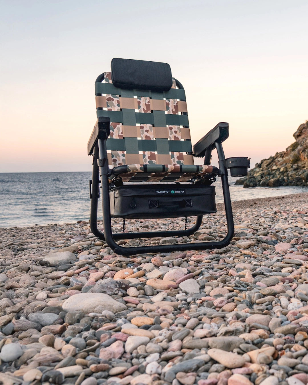 One PARKIT x Free Fly Eclipse Recliner chairs in the limited-edition Vintage Camo pattern sit on a rocky beach at sunset, facing the ocean and rugged coastal cliffs, showcasing premium outdoor design, durable materials, and the adventure-ready comfort of PARKIT’s 3-in-1 recliner chair built for life outside in mobile format.