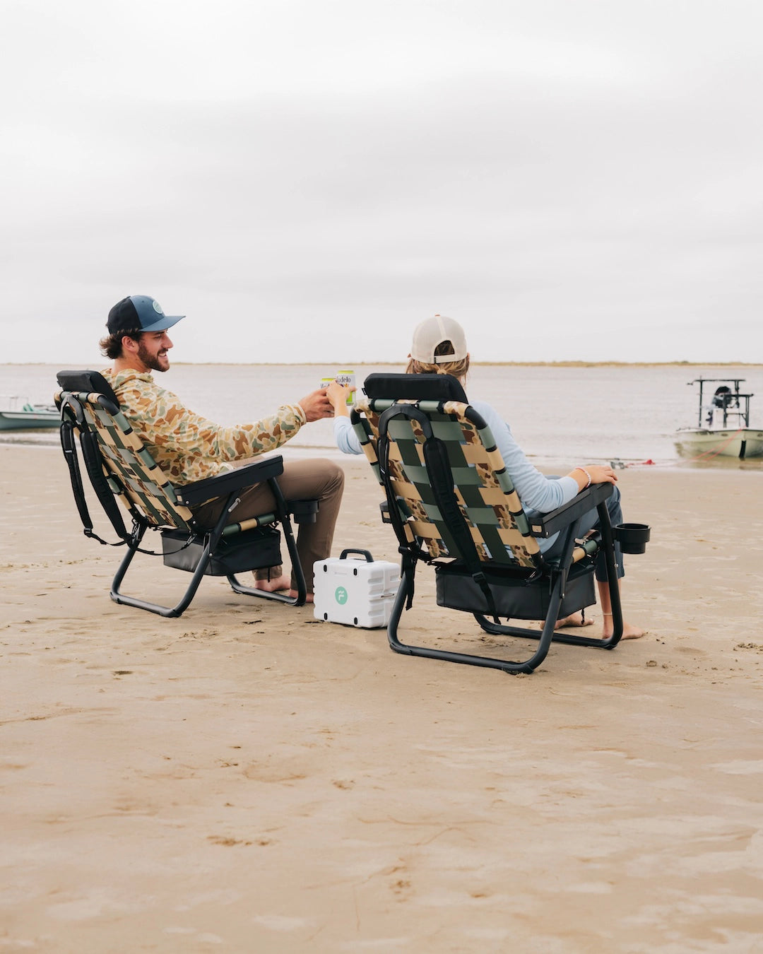 Two people relaxing on a sandy beach in PARKIT x Free Fly Eclipse Recliner chairs in Vintage Camo pattern, toasting drinks beside a white Turtle box speaker while a fishing skiff rests in shallow water capturing coastal adventure, outdoor connection, and premium 3-in-1 chair design built for life by the water in mobile format.