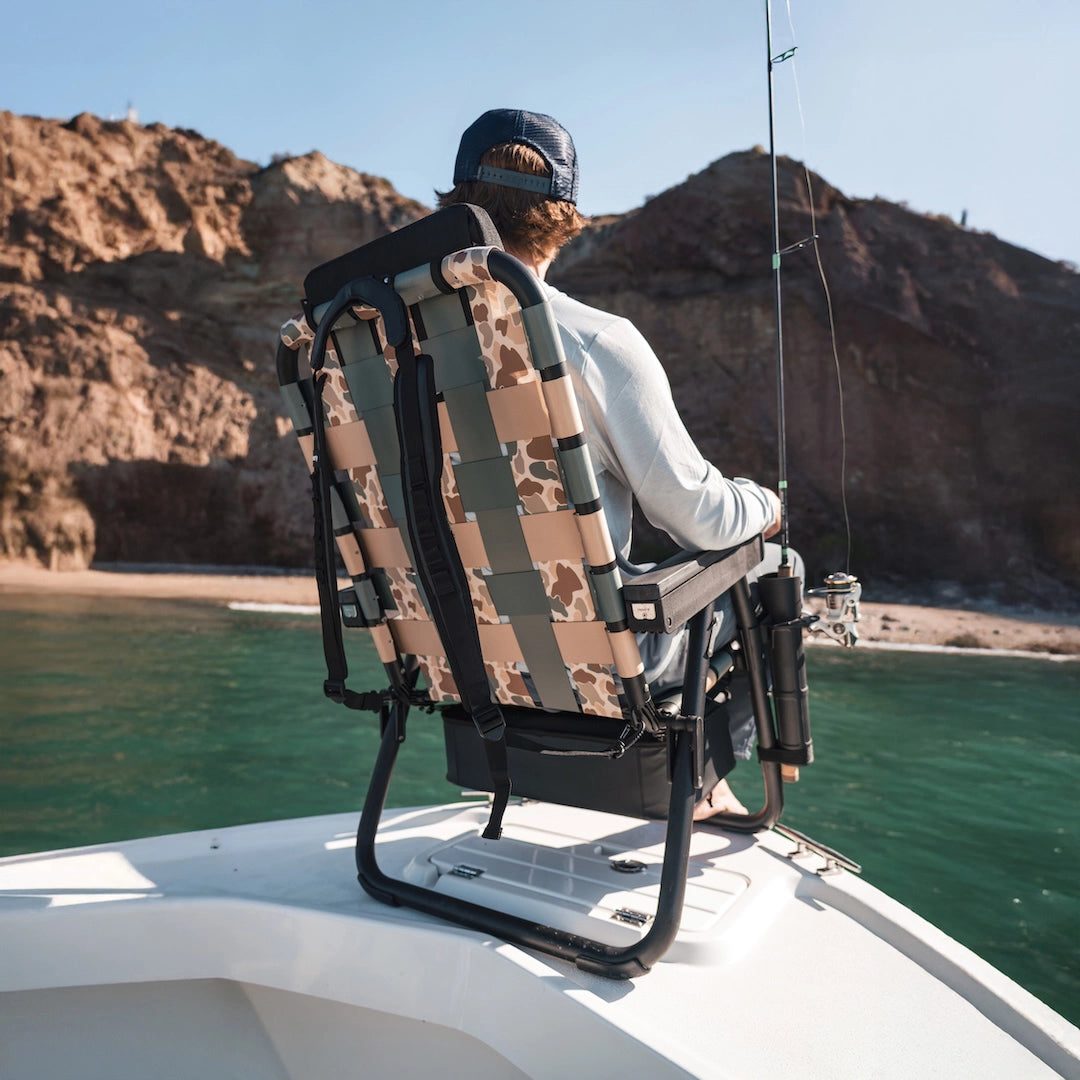 Man fishing from a boat while sitting in the PARKIT × Free Fly Eclipse Recliner in Vintage Camo, featuring the Outpost accessory mount holding a fishing rod. This premium folding outdoor recliner combines comfort and function with backpack carry straps, built-in cooler, and marine-grade materials — the perfect fishing chair for dads, outdoor adventurers, and boat lovers seeking durability and all-day relaxation.
