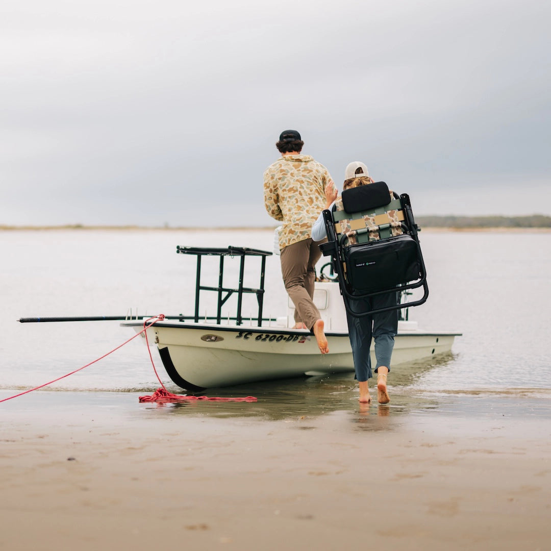 Two people walking barefoot toward a skiff, one carrying the PARKIT × Free Fly Eclipse Recliner in Vintage Camo using its backpack carry straps. The folding outdoor chair features an integrated cooler and rugged aluminum frame the ultimate portable beach, boat, and camping chair for couples, adventurers, and weekend explorers.