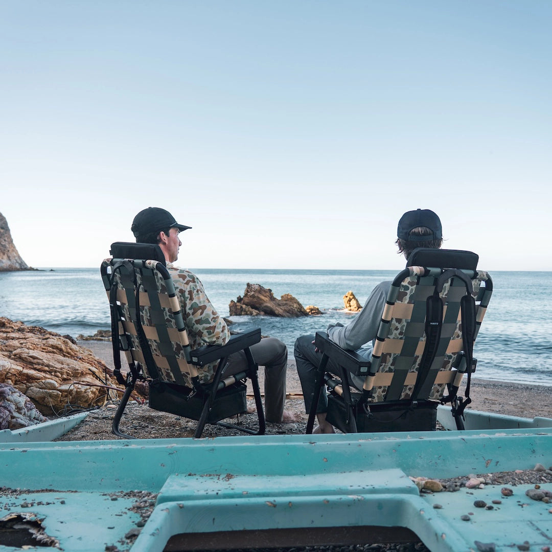 Two friends relaxing on Catalina Island in PARKIT × Free Fly Eclipse Recliners in Vintage Camo, overlooking the Pacific Ocean. The rugged folding outdoor chairs feature backpack straps, a built-in cooler, and marine-grade materials ideal for beach camping, coastal adventures, and the best gift for dads or moms who love to unwind outdoors.