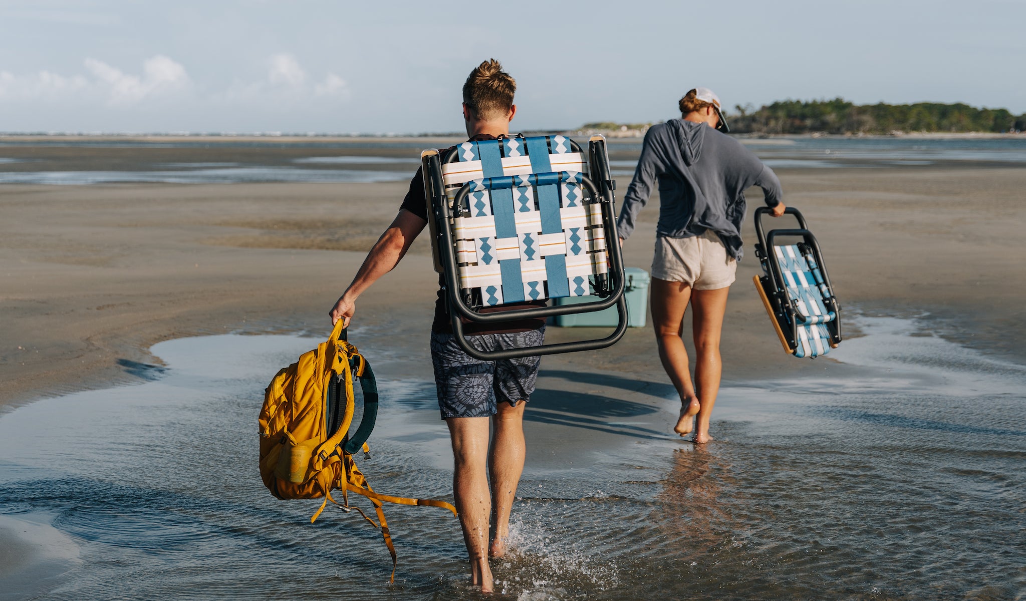 A boy and a girl carrying their Voyager Outdoor chairs to a sandbar party and campout in the marshlands outside of Charleston, South Carolina