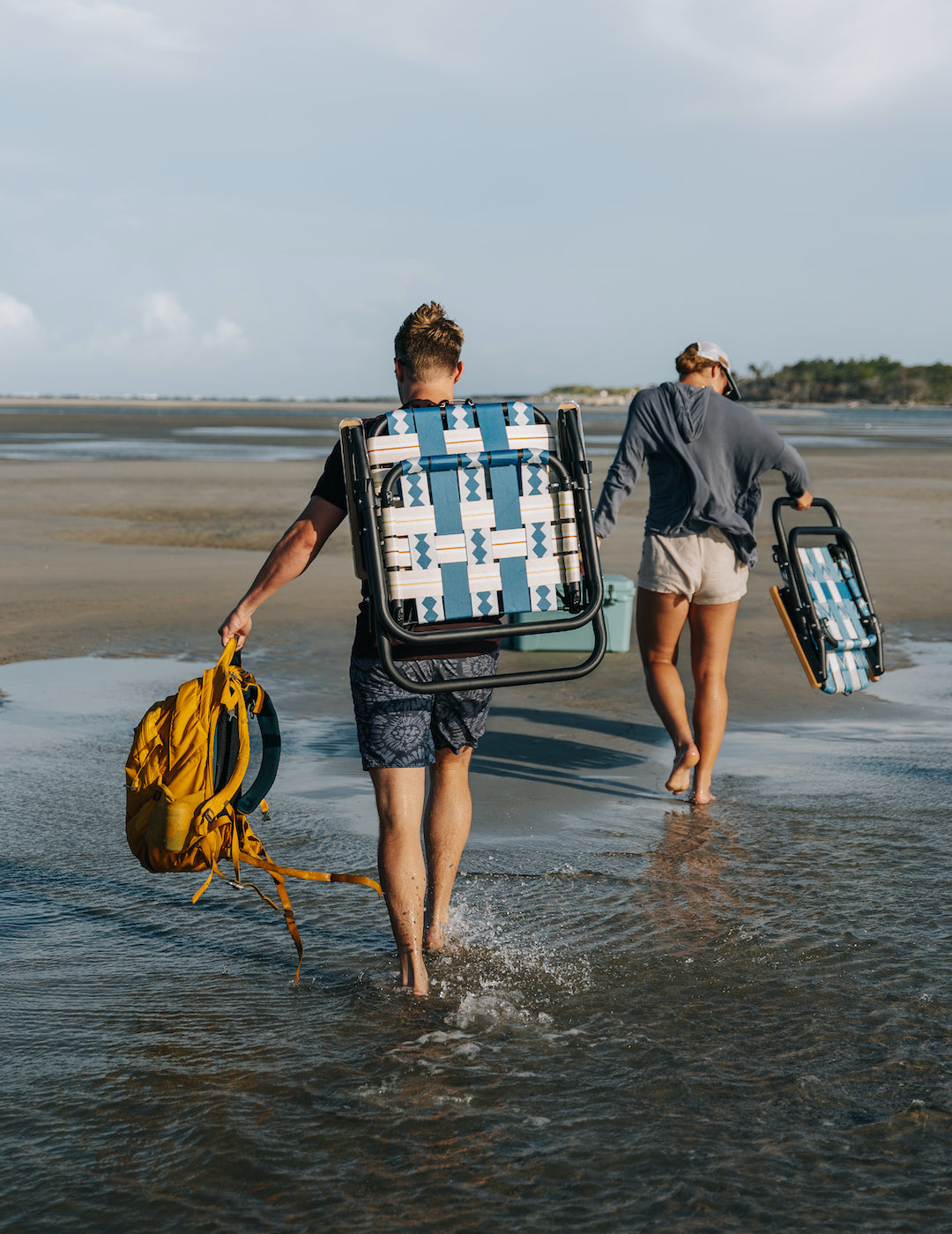 A boy and a girl carrying their Voyager Outdoor chairs to a sandbar party and campout in the marshlands outside of Charleston, South Carolina
