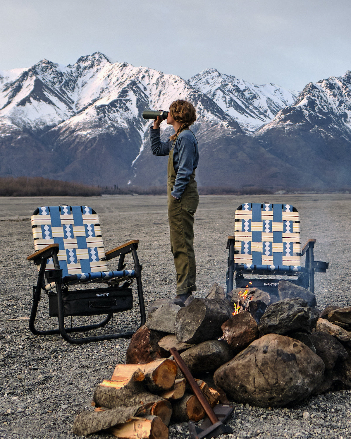 A woman in Alaska with her two PARKIT Voyager Camping chairs and a fire nestled in front of massive snowy mountains. 