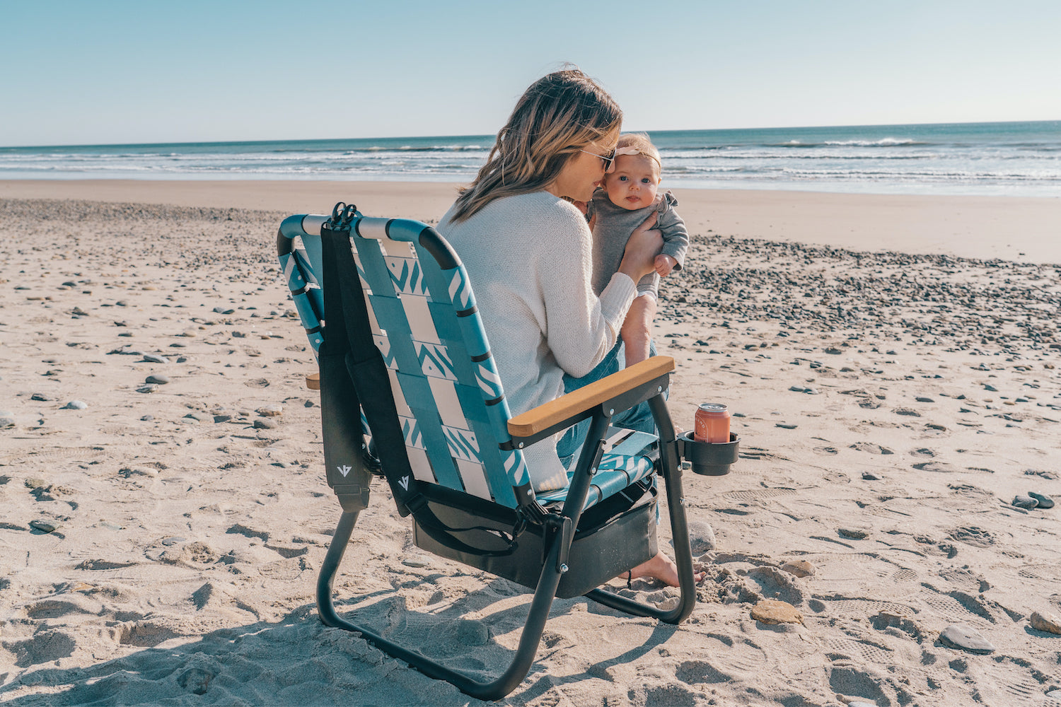 A Mom sitting in the Voyager outdoor chair at the beach while holding her baby girl and drinking an ashland seltzer in the cupholder while waves break in the background.