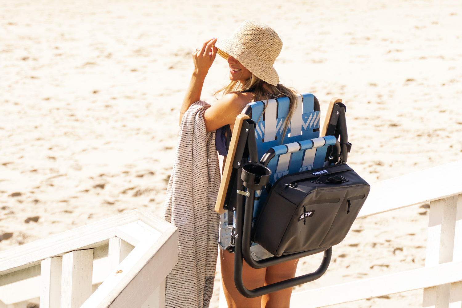 A woman carrying a voyager outdoor chair in glacier down to the beach while wearing a sun hat. She's carrying the voyager outdoor chair like a backpack with a full cooler from PARKIT.