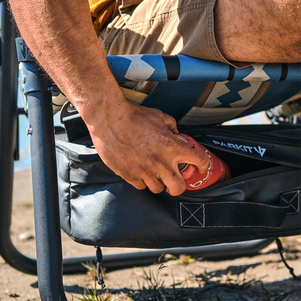 A Man pulling a beverage or drink out of his parkit voyager chair cooler while sitting at a campsite.