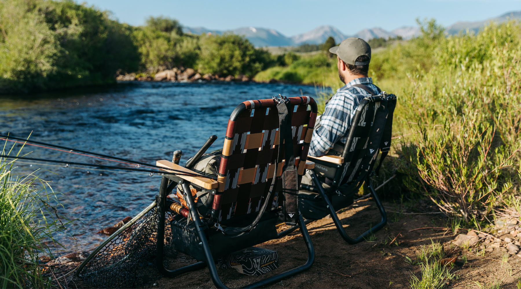 A man sitting alongside the river with his Voyager outdoor chairs while he and his friend wait for the fish to bite
