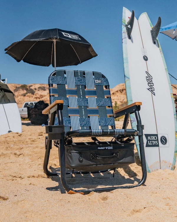 PARKIT voyager beach chair built in collaboration with salty crew set up on the beach surrounded by surfboards and beach day gear like umbrellas and fishing gear. 