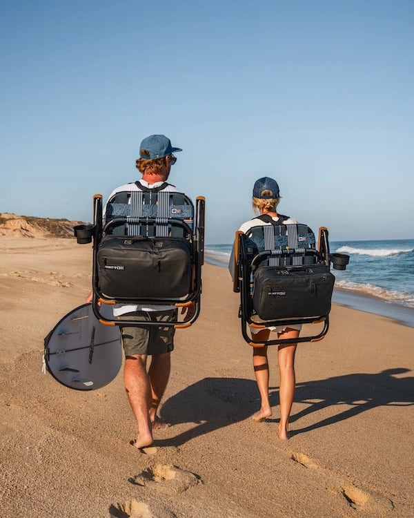 a couple carrying their parkit and salty crew collaboration chairs hands free  while walking down the coast looking for the best spot to set up camp. 