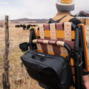 Person with a patterned chair and black bag in a field with cows in the background
