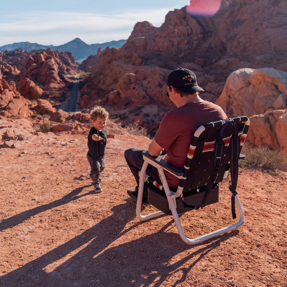 Man and child in a desert landscape with mountains in the background sitting in ia black and white folding chair with red stripes