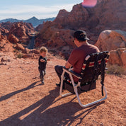 Man and child in a desert landscape with mountains in the background sitting in ia black and white folding chair with red stripes