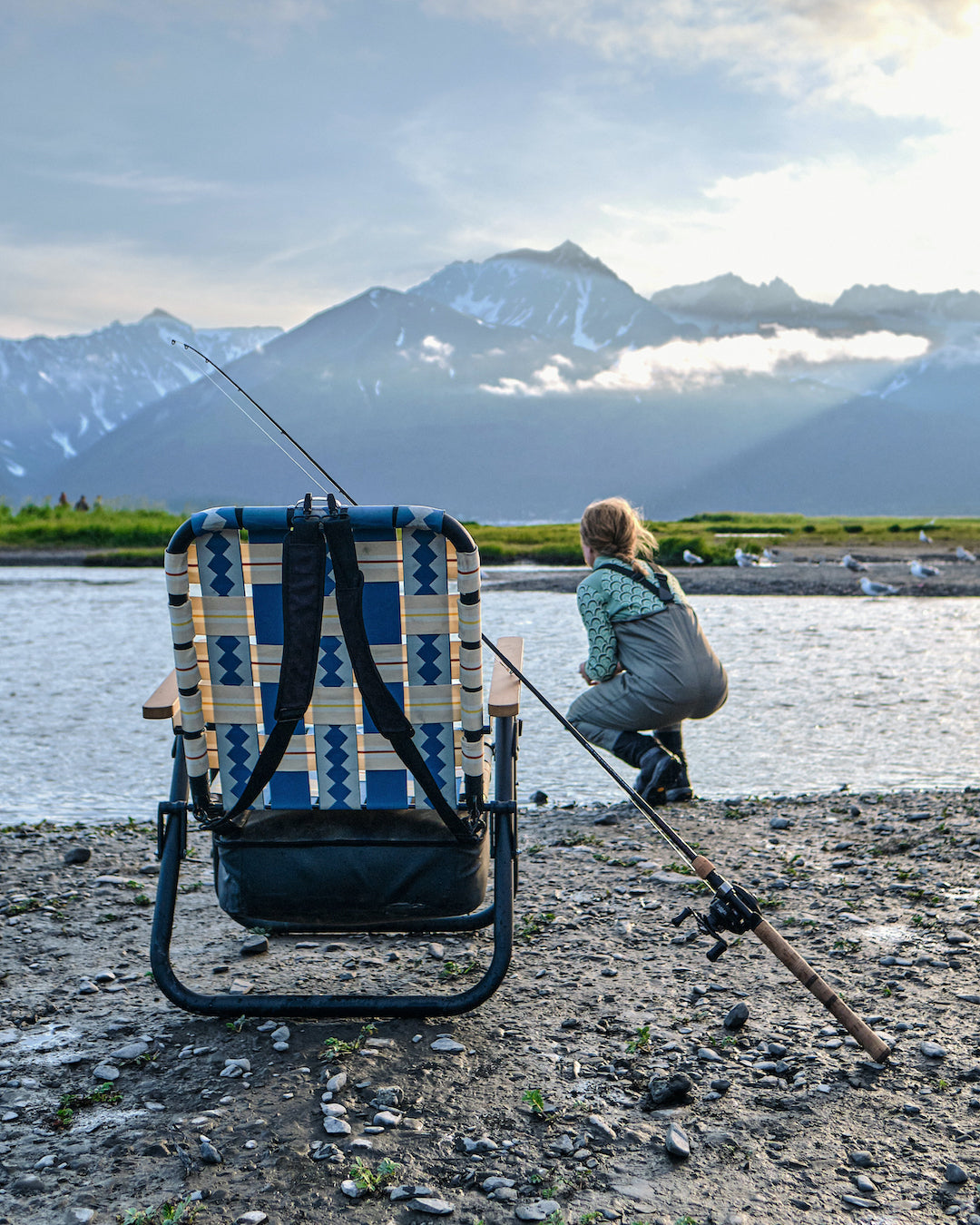 A woman fishing in Alaska with the PARKIT Voyager outdoor chair sitting on the river bank which holds all of her fishing supplies in the attachable cooler.