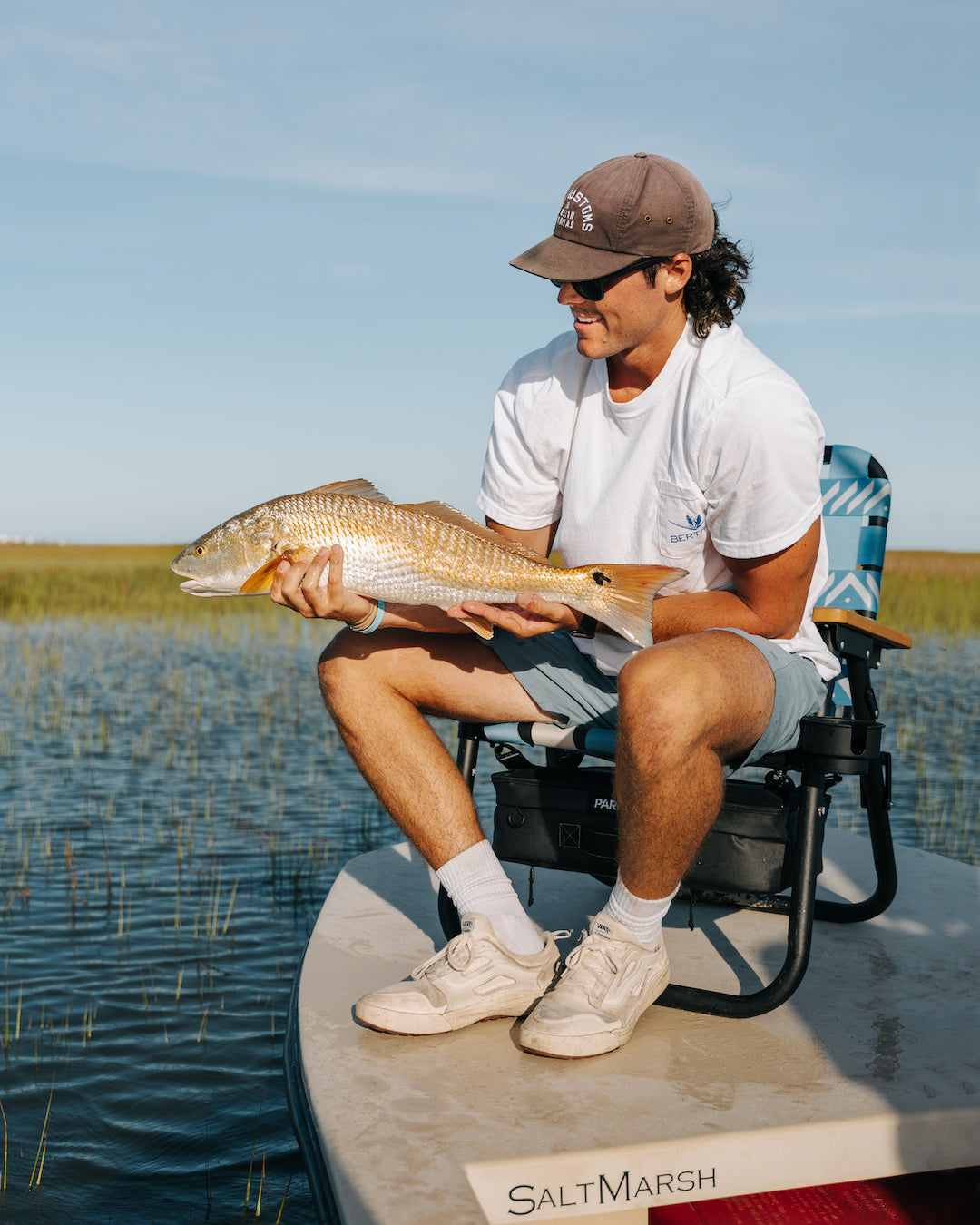 A man sitting in his PARKIT Voyager Outdoor Chair with the best fish he caught all day. He is sitting on the bow of a skiff in the PARKIT Voyager Chair in Glacier Blue
