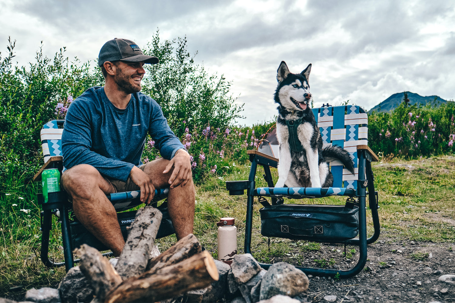 A man and his dog sit around a campfire in their Classic Blue Voyager outdoor chairs from PARKIT.