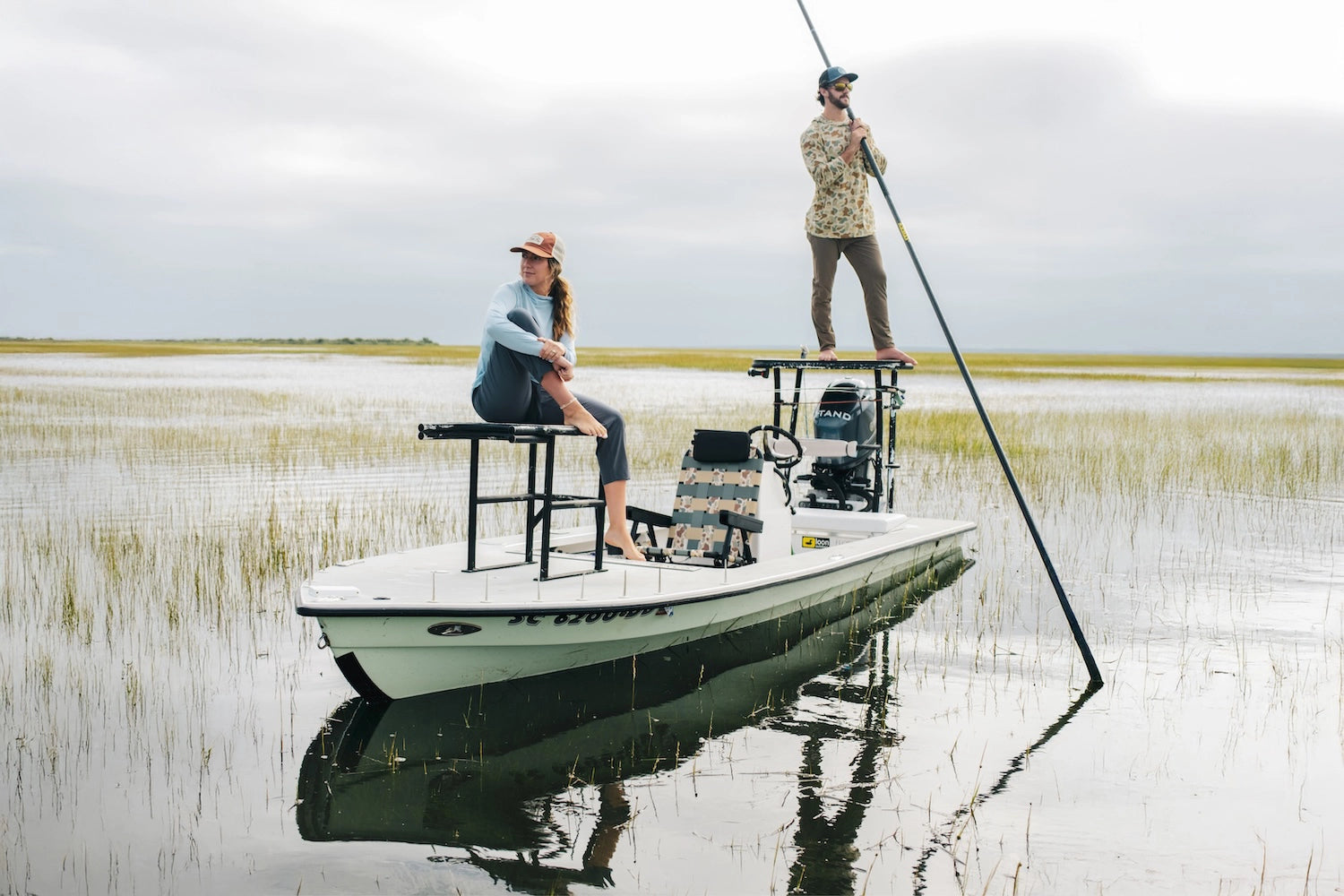 Two anglers on a flats skiff surrounded by coastal marsh, featuring the PARKIT × Free Fly Eclipse Recliner in Vintage Camo pattern, capturing the spirit of adventure, premium outdoor gear, and the lifestyle connection between performance apparel and purpose-built comfort on the water.