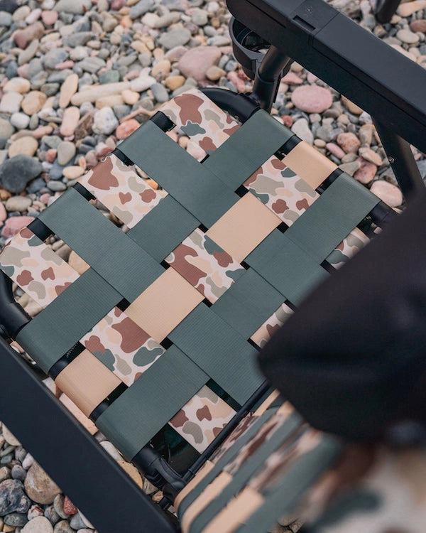 Close-up of the PARKIT × Free Fly Eclipse Recliner seat showing the Vintage Camo woven webbing pattern that blends tan, green, and brown tones against a bed of river rocks, highlighting the durable outdoor materials, craftsmanship, and camouflage aesthetic inspired by natural landscapes.