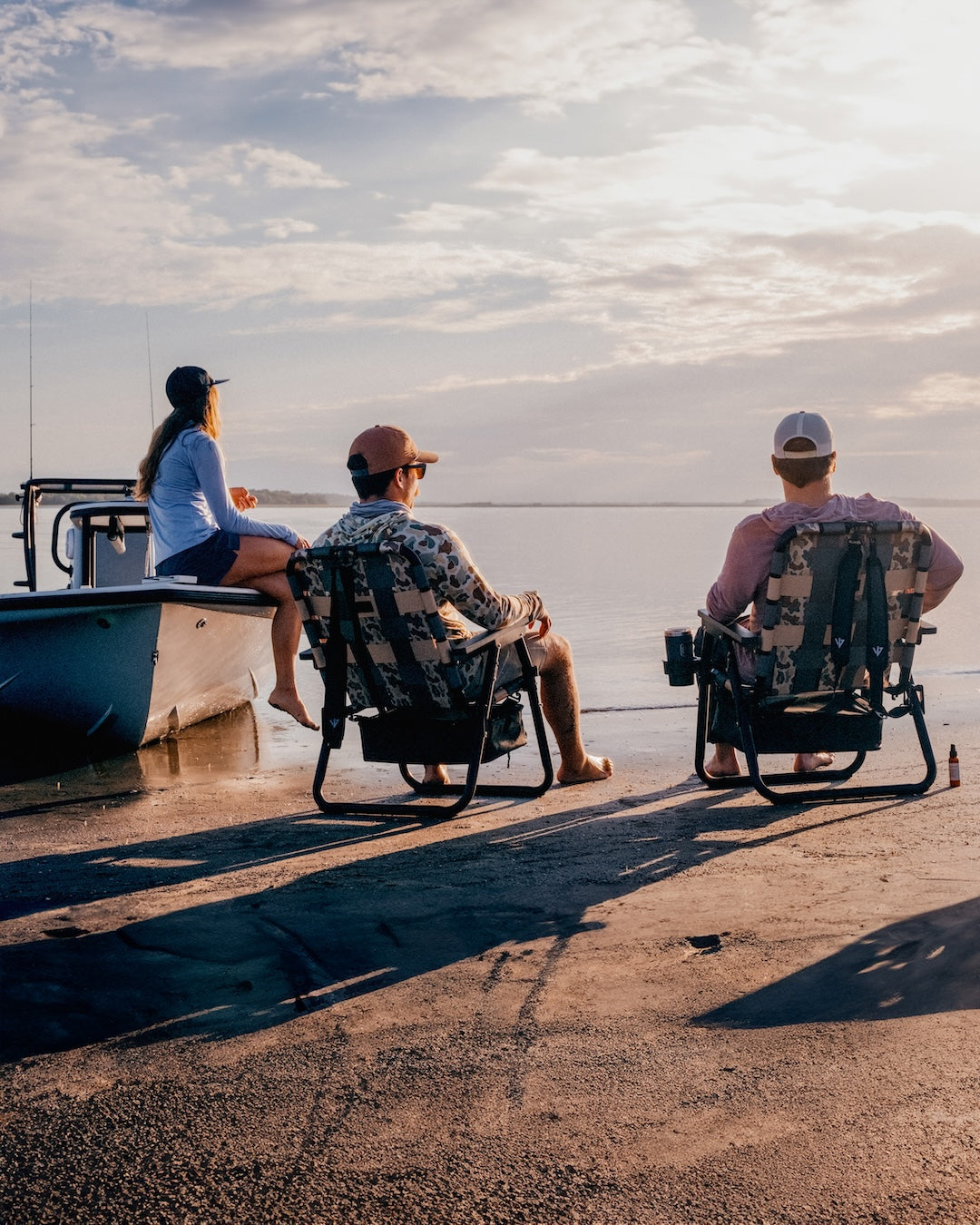 a group of friends sitting on the shore of a beach, one on a boat and two on beach chairs 