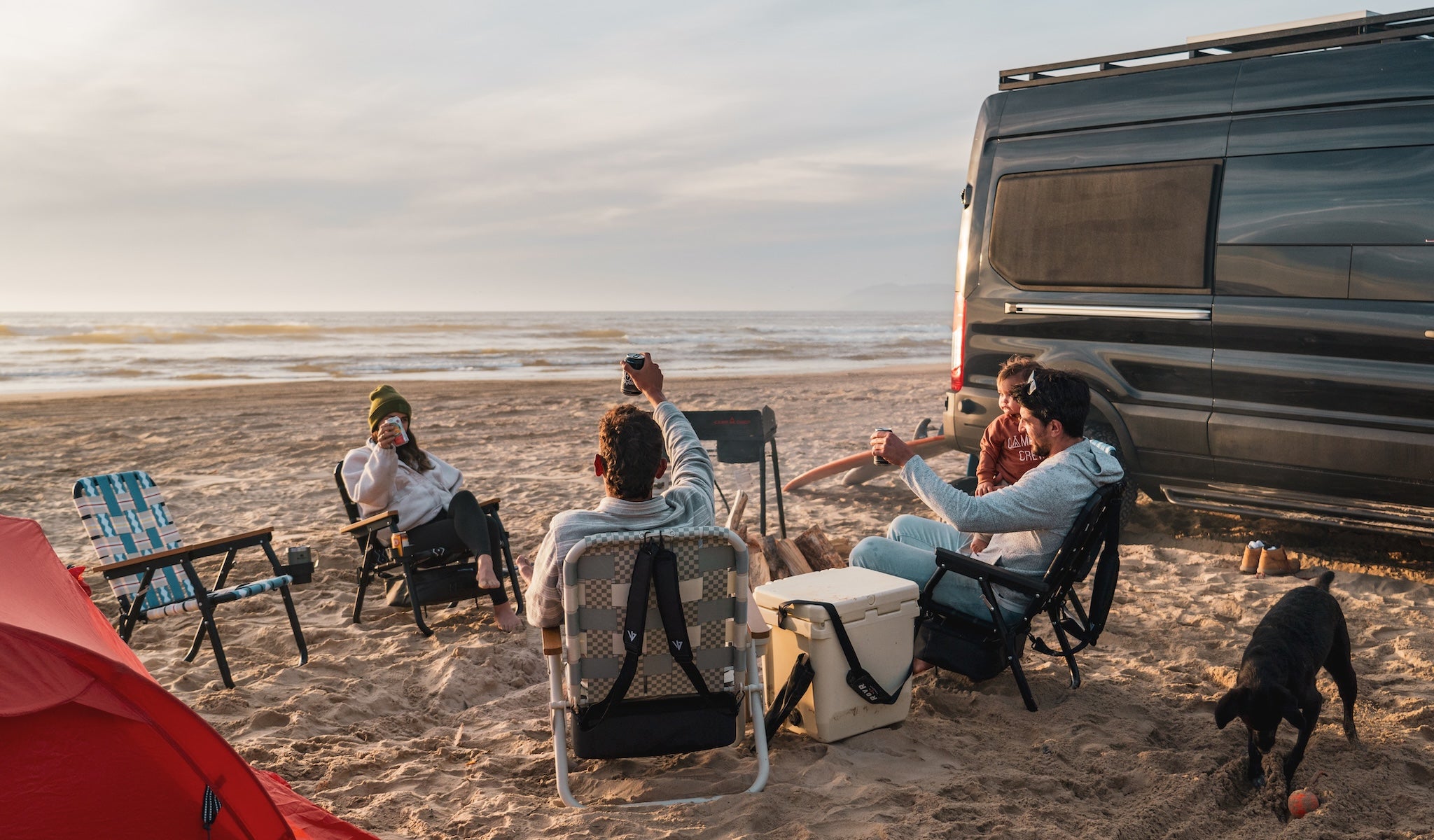 Friends sitting in PARKIT Voyager Campaing chairs at their campsite in Pismo Beach where their camper van is parked on the sand and the sun sets with crashing waves in the background