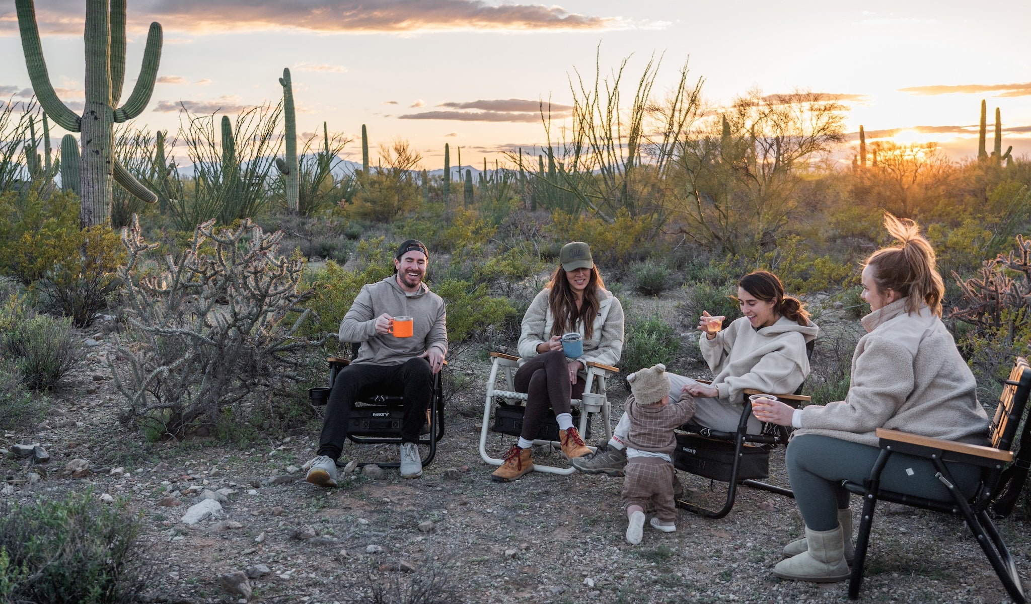 Group of friends and family sitting in PARKIT Voyager outdoor chairs around a desert campsite at sunset, drinking coffee and relaxing among cacti, showcasing durable adventure-built chairs designed for comfort, connection, and life outside.